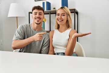 Poster - Young beautiful couple wearing casual clothes sitting on the table at home amazed and smiling to the camera while presenting with hand and pointing with finger.