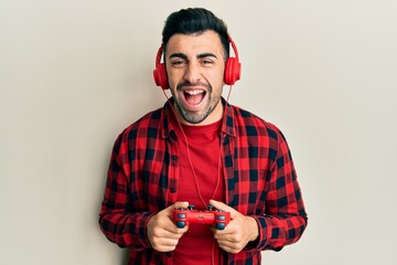 Young hispanic man playing video game holding controller smiling and laughing hard out loud because funny crazy joke.