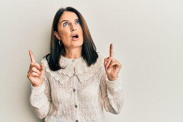 Middle age brunette woman wearing casual clothes amazed and surprised looking up and pointing with fingers and raised arms.