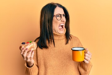 Canvas Print - Middle age brunette woman eating pastry and drinking coffee angry and mad screaming frustrated and furious, shouting with anger. rage and aggressive concept.