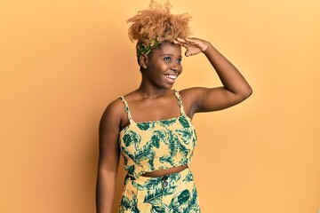 Poster - Young african woman with afro hair wearing summer dress very happy and smiling looking far away with hand over head. searching concept.