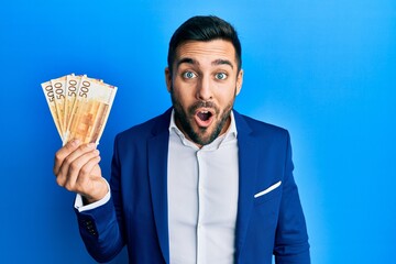 Canvas Print - Young hispanic businessman wearing business suit holding norwegian krone banknotes scared and amazed with open mouth for surprise, disbelief face