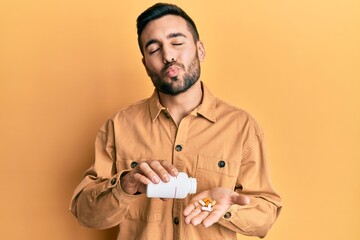 Poster - Young hispanic man holding pills looking at the camera blowing a kiss being lovely and sexy. love expression.