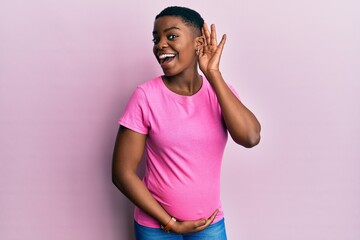 Wall Mural - Young african american woman expecting a baby, touching pregnant belly smiling with hand over ear listening an hearing to rumor or gossip. deafness concept.