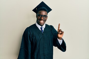 Wall Mural - Handsome black man wearing graduation cap and ceremony robe pointing finger up with successful idea. exited and happy. number one.