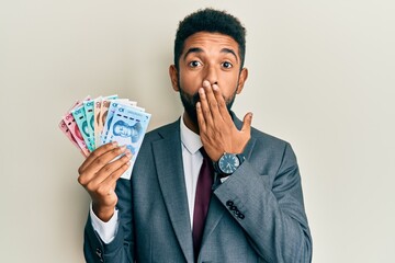 Poster - Handsome hispanic man with beard wearing business suit holding yuan banknotes covering mouth with hand, shocked and afraid for mistake. surprised expression