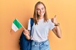 Beautiful blonde woman exchange student holding ireland flag smiling happy and positive, thumb up doing excellent and approval sign