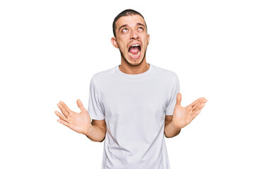 Hispanic young man wearing casual white t shirt crazy and mad shouting and yelling with aggressive expression and arms raised. frustration concept.