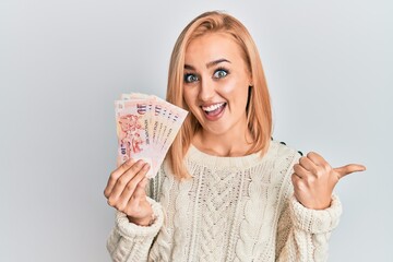 Beautiful caucasian woman holding 10 singapore dollars banknotes pointing thumb up to the side smiling happy with open mouth