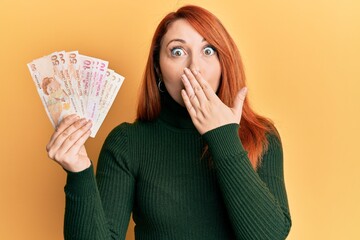 Poster - Beautiful redhead woman holding turkish lira banknotes covering mouth with hand, shocked and afraid for mistake. surprised expression