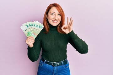 Poster - Beautiful redhead woman holding 50 hong kong dollars banknotes doing ok sign with fingers, smiling friendly gesturing excellent symbol