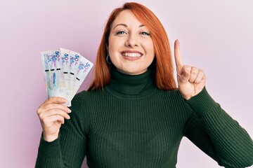 Canvas Print - Beautiful redhead woman holding 50 colombian pesos banknotes smiling with an idea or question pointing finger with happy face, number one