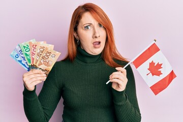 Canvas Print - Beautiful redhead woman holding canadian dollars and canada flag in shock face, looking skeptical and sarcastic, surprised with open mouth