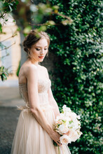 Pensive Bride In Pink Dress With A Bouquet Of Flowers Bowed Her Head In A Background Of Green Plants. Lake Como