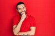 © Krakenimages.com - Hispanic man with beard wearing casual red t shirt with hand on chin thinking about question, pensive expression. smiling with thoughtful face. doubt concept.