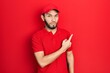 © Krakenimages.com - Hispanic man with beard wearing delivery uniform and cap pointing with hand finger to the side showing advertisement, serious and calm face