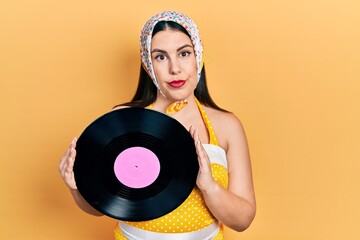 Poster - Young hispanic woman wearing pin up style holding vinyl disc relaxed with serious expression on face. simple and natural looking at the camera.