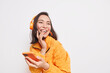 © wayhome.studio  - Glad Asian woman laughs happlily looks away into distance enjoys favorite music playlist uses mobile application wears wireless headphones dressed in orange jacket isolated on white wall blank space