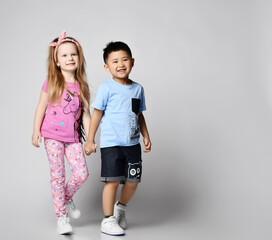 Two happy laughing, smiling kids friends Asian boy and Caucasian girl in summer clothes have good time together, walking coming to us holding hands over light background with copy space