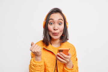 Wall Mural - Photo of good looking Asian woman with dark hair clenches fist uses smartphone for online communication gets new music subscription dressed in orange jacket isolated over white studio background