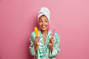 Wall Mural - Domestic style and summer dessert concept. Positive dark skinned woman smiles happily holds two delicious ice creams on sticks wears silk pajama wrapped bath towel on head has happy mood poses indoor