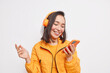 © wayhome.studio  - Modern gadget and lifestyle concept. Positive Asian woman concentrated into smartphone display enjoys listening music from playlist wears orange jacket smiles cheerfully isolated over white background