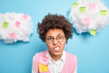 Poster - Close up shot of irritated curly haired young female office worker clenches teeth looks with annoyed expression has deadline and much tasks writes down information to remember on memo stickers