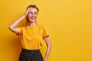 Horizontal shot of pretty joyful woman looks away keeps hand on head smiles broadly wears basic t shirt and skirt optical eyeglasses isolated over yellow background blank space for your advert