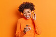 Photo of cheerful African American woman chooses song to listen from her playlist enjoys good sound in headphones concentrated in smartphone display dressed casually isolated over orange wall