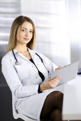 Wall Mural - Young optimistic woman-doctor is using a clipboard, while sitting at the desk in her cabinet in a clinic. Portrait of friendly female physician woman with a stethoscope. Perfect medical service in a