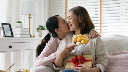 Attractive beautiful asian middle age mum sit with grown up daughter give gift box and flower in family moment celebrate mother day. Overjoy bonding cheerful kid embrace relationship with retired mom.