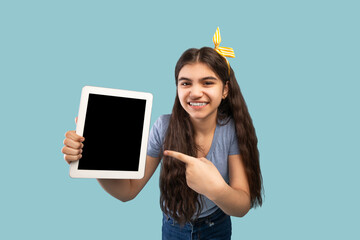 Smiling Indian teenage girl pointing at tablet with white blank screen on blue background, space for website or app