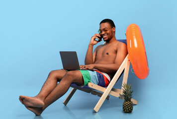 Online work on summer vacation. Black guy with laptop speaking on smartphone while sitting in lounge chair