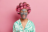 Fototapeta  - Depressed sad woman cries loudly has mournful expression applies beauty mask on face hair rollers prepares for date upset to have breaking with husband dressed casually isolated on pink wall