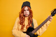 © WHstudio Leushin N - Distressed unhappy redhead young woman plays bass electric guitar has sad expression wears black hat and casual yellow sweatshirt poses indoor. Displeased female rocker with musical instrument
