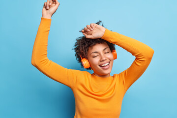 Wall Mural - Photo of relaxed curly haired young woman keeps eyes closed dances and catches every bit of music smiles broadly wears casual orange jumper uses wireless headphones isolated over blue background