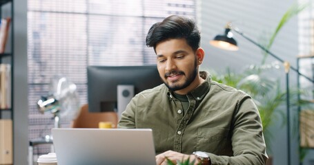 Close up portrait of cheerful Hindu young handsome guy worker videochatting speaking on online call on laptop while sitting at workplace at office and smiling waving hand. Successful businessman