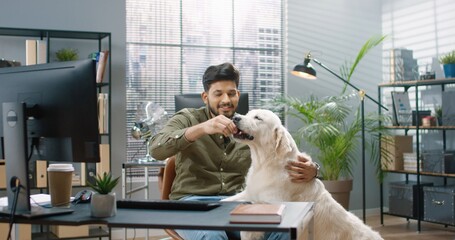 Portrait of happy joyful Hindu male sitting in cabinet at work feeding his cute dog pet in good mood. Handsome cheerful bearded guy at office train and feed animal. Positive emotions, pet concept