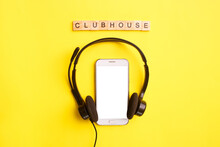 Headphones And Smartphone With Wooden Letters On A Yellow Background. Clubhouse Social Media Concept. Mock Up, Copy Space, Flat Lay, Top View