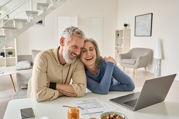 Happy mature older family couple laughing, bonding sitting at home table with laptop. Smiling middle aged senior 50s husband and wife having fun satisfied with buying insurance, paying bills online.