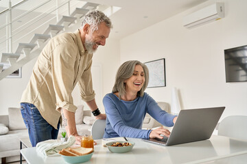 Happy middle aged 50s family couple having fun using laptop computer technology at home. Smiling senior older mature husband and wife laughing shopping online or having virtual meeting in living room.