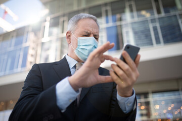 Wall Mural - Covid concept, masked elder businessman using his smartphone outdoor