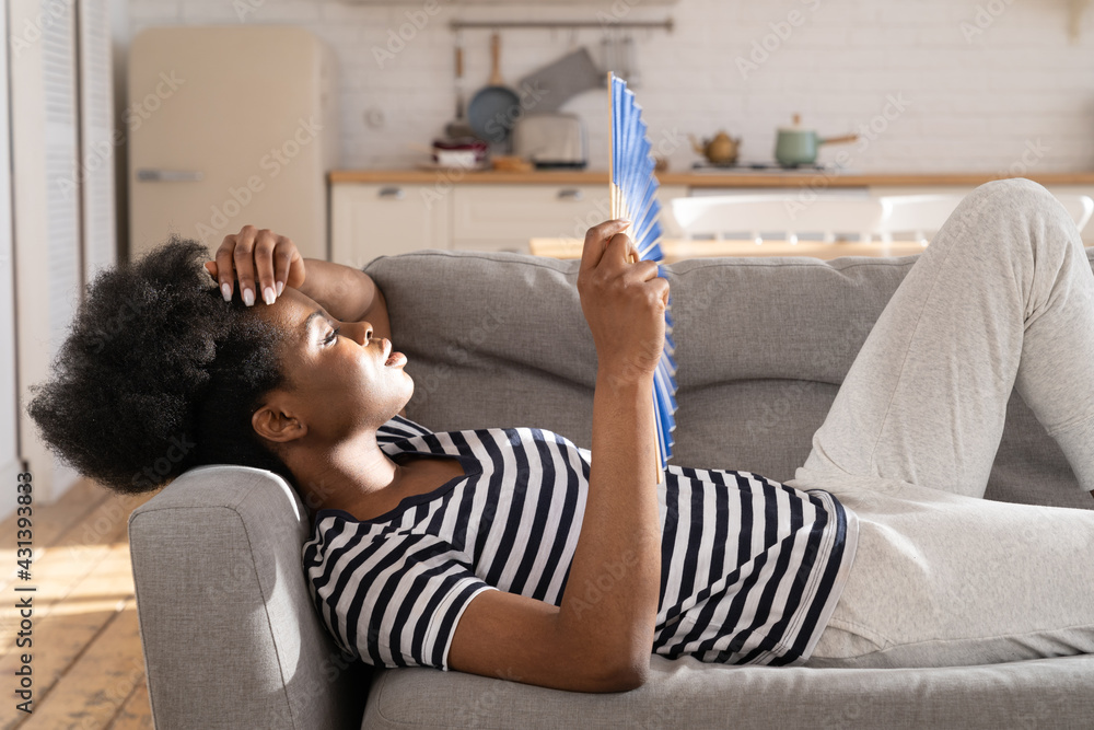 Exhausted overheated black female lying on sofa with paper fan suffer from hot temperature inside and headache. Shocked african woman breathe fresh cooled air to calm down panic attack alone at home