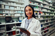 © StratfordProductions - Happy woman working in pharmacy using digital tablet wearing labcoat looking at camera