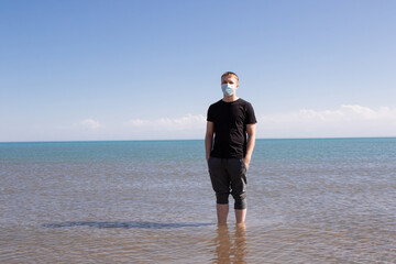 Wall Mural - a guy in a blue medical mask stands in the sea. Rest during quarantine.