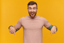 Cheerful Attractive Young Bearded Man With Tattoo On Hand In Pink Tshirt Looks Excited And Pointing Down By Fingers Of Both Hands Over Yellow Background