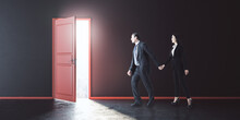 Businessman And Businesswoman Walking Towards An Exit In A Dark Concrete Room With A Red Door. Leadership And Future Concept