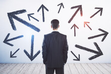 Businessman Standing Puzzled In Front Of A Wall With Multiple Black Arrows Pointed In Different Directions. Decision Making And Career Path Concept