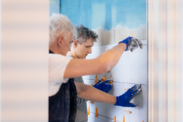 Two tilers installing wall tile at home. Father and son laying tiles on a bathroom wall. DIY, Do it yourself concept. House improvement. Repair renovation work. Selective focus, copy space