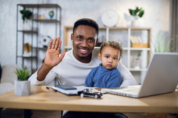 Wall Mural - Smiling african man waving on camera while sitting at home office with his cute baby boy on knees. Concept of parenting, technology and freelance.